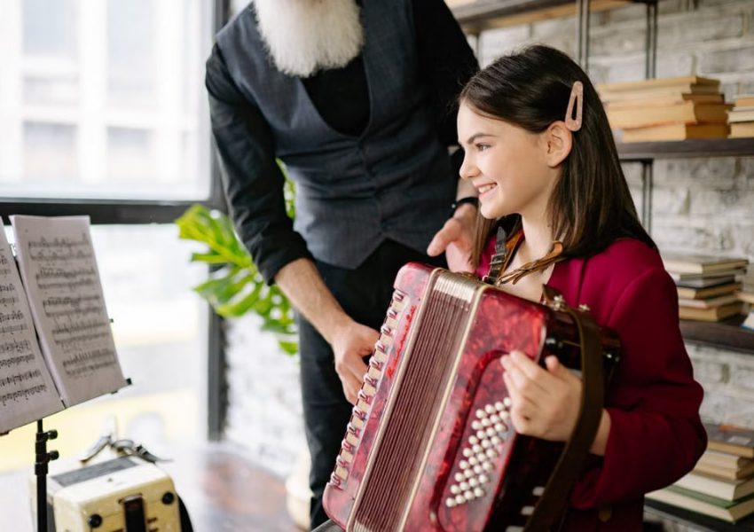 A young girl learning accordion indoors, guided by an adult musician.