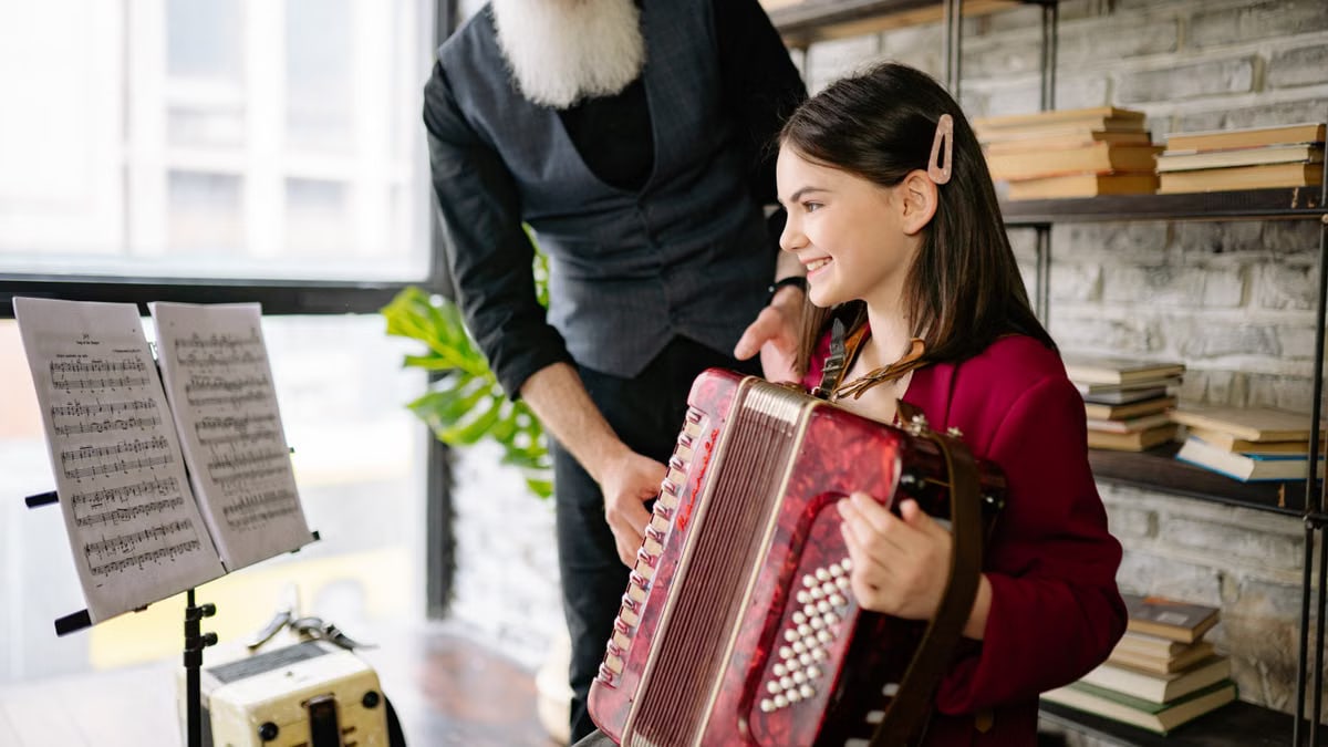 A young girl learning accordion indoors, guided by an adult musician.