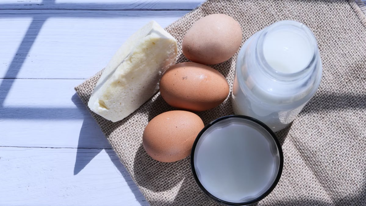 Overhead view of eggs, milk, and cheese on burlap, highlighting rustic food essentials.